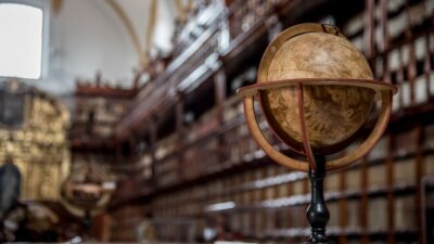 Featured image for Is Dark Academia Still Popular? to illustrate the dark academia aesthetic, it shows a picture of an antique library in the background with an old globe in focus on a table. The picture's vibe is dark and moody.