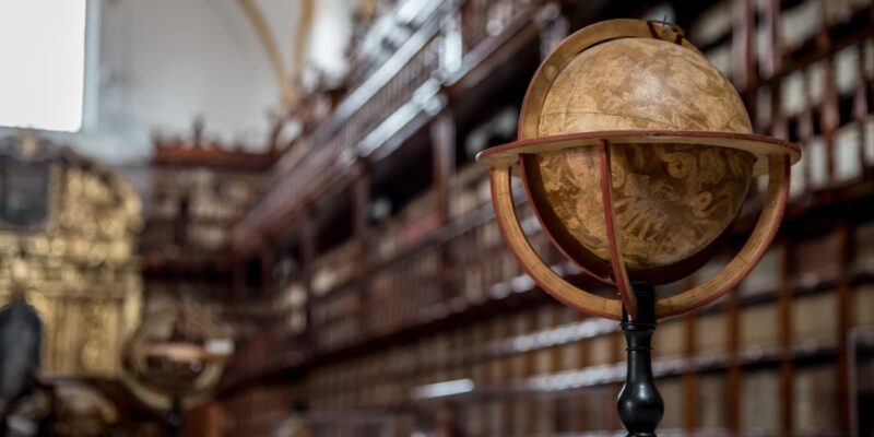 Featured image for Is Dark Academia Still Popular? to illustrate the dark academia aesthetic, it shows a picture of an antique library in the background with an old globe in focus on a table. The picture's vibe is dark and moody.